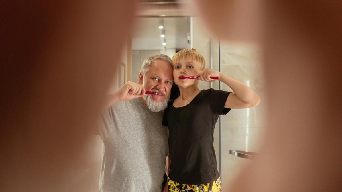 Family Cosmetic Dentistry Worcester: One Dental Home for Every Smile - Friendly Dental of Worcester Father and son brushing teeth together in a bathroom, pink toothbrushes touching teeth, smiling at the mirror through a blurred foreground frame