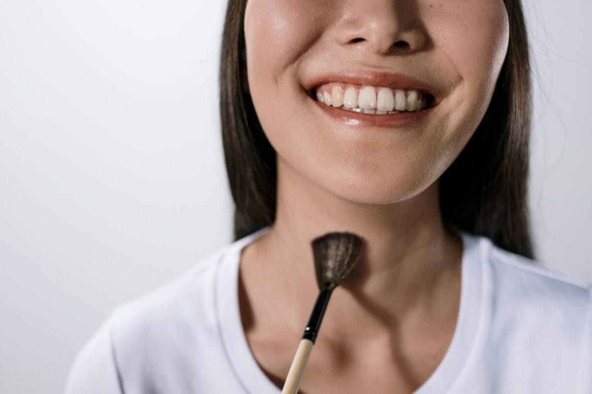 Family Cosmetic Dentistry Worcester: Keeping Smiles Youthful - Friendly Dental of Worcester Close-up of a smiling woman's mouth and chin with a makeup brush resting near her neck.