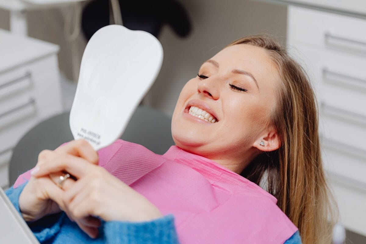 Woman in a dental chair wearing a pink bib, smiling while looking at a handheld mirror in a clinic setting.
