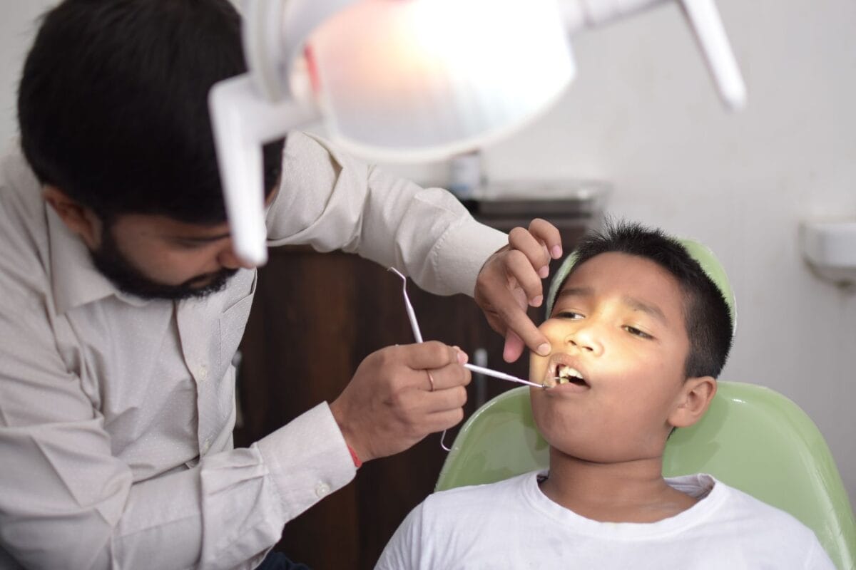 Dentist examining a boy's mouth with dental instruments in a clinic, boy seated in green chair.