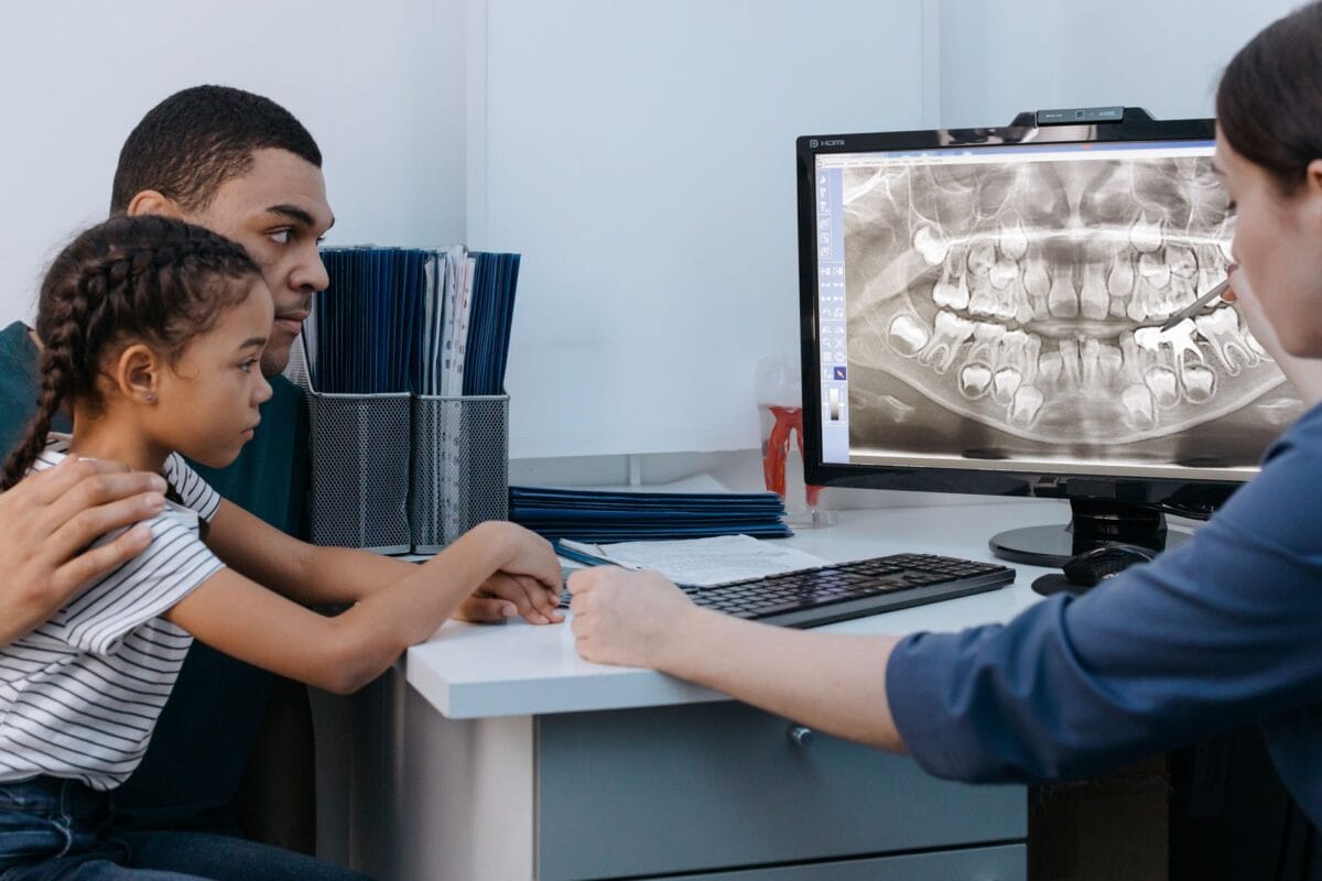 Dentist points at a dental X-ray on a monitor as a father and daughter review it at the desk in a clinic office.