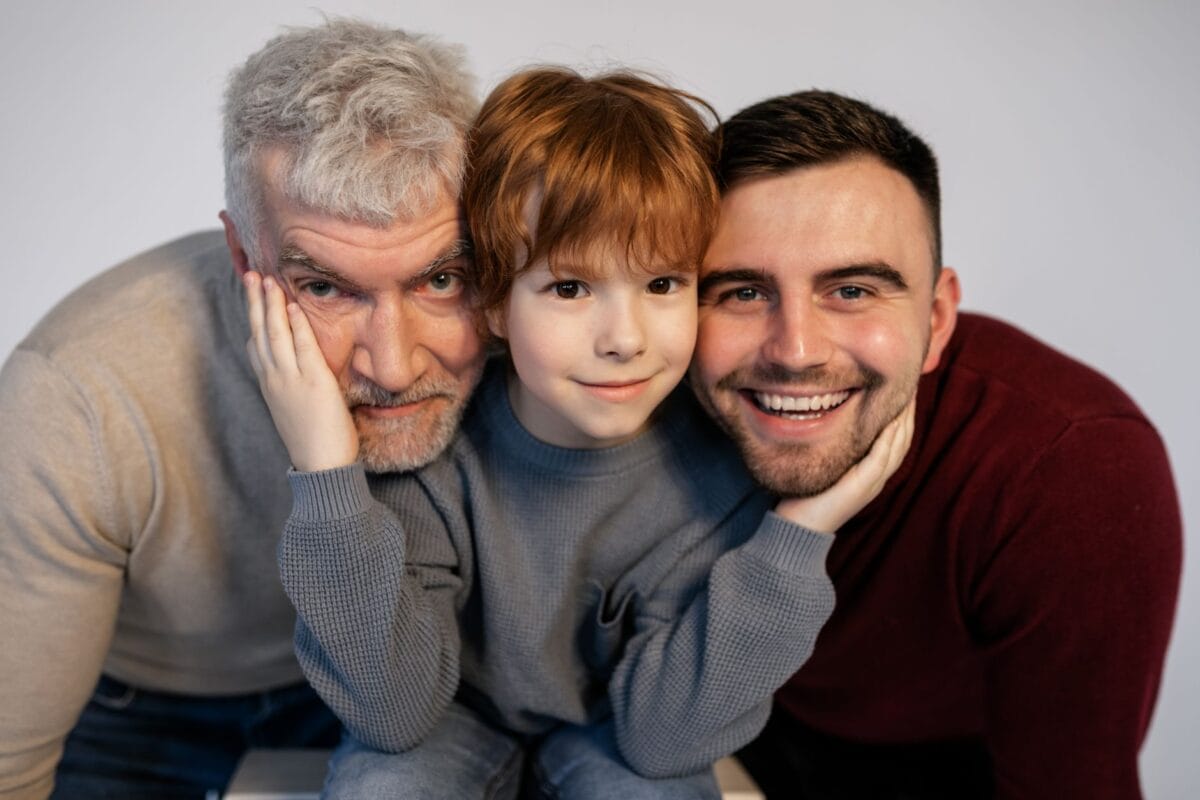 Three generations—grandfather, child, and father—smile warmly as they lean their heads together for a family portrait.