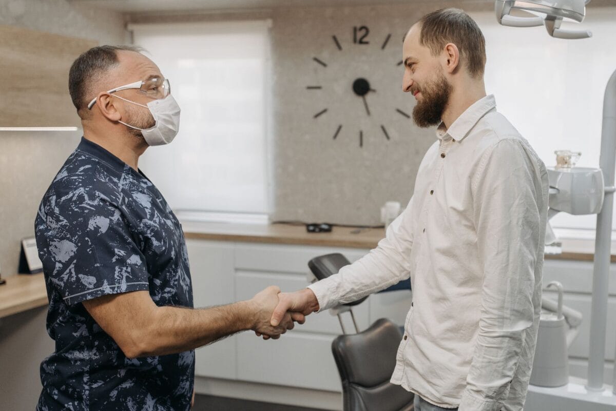 Dentist wearing a mask and protective glasses shakes hands with a patient in a dental clinic with a chair in the background.