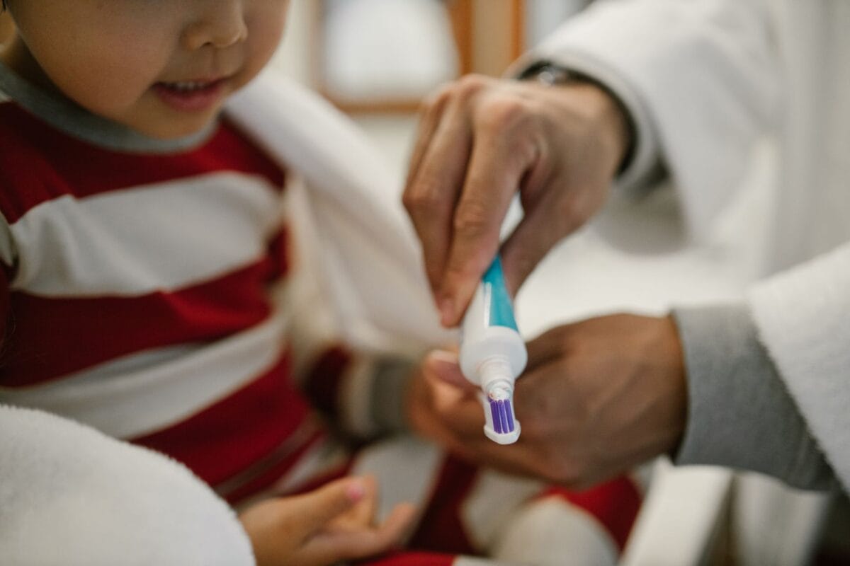 Child wearing a red and white striped shirt brushing teeth with an adult guiding the toothbrush nearby in a bright setting.